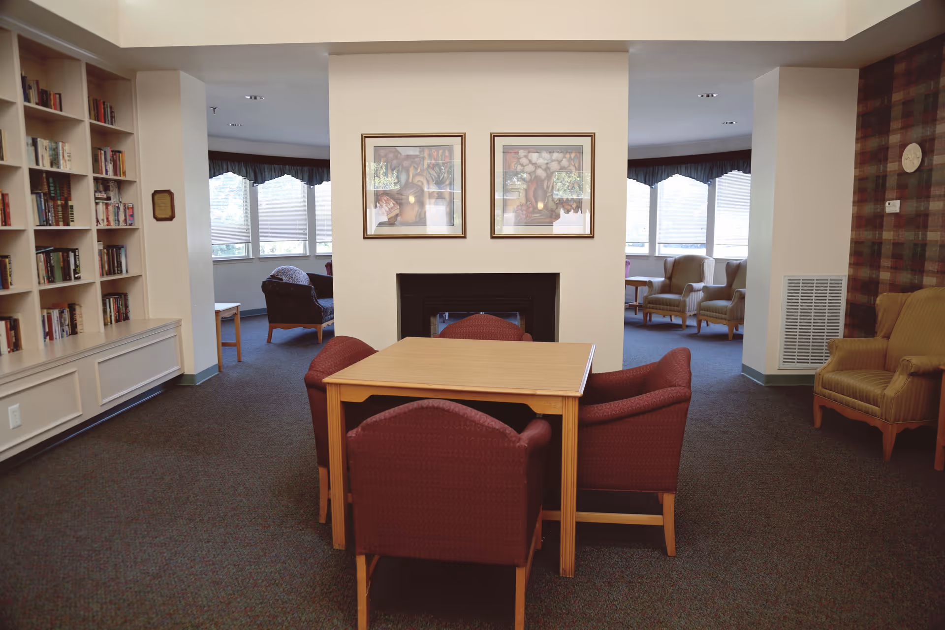 A cozy senior living community common area with a wooden table surrounded by four red upholstered chairs in the center. Behind the table is a fireplace with two framed pictures hanging above it. To the left is a built-in bookshelf filled with books, and in the background, there are additional seating areas with armchairs near large windows letting in natural light.