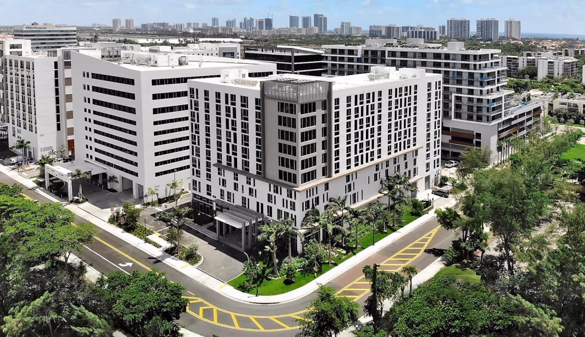 Aerial view of a large multi-story white senior living building with surrounding roads, landscaping, and nearby high-rise buildings.