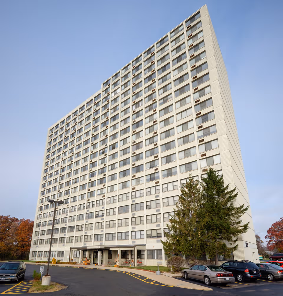 Tall white high-rise senior residence building with a parking lot and trees.