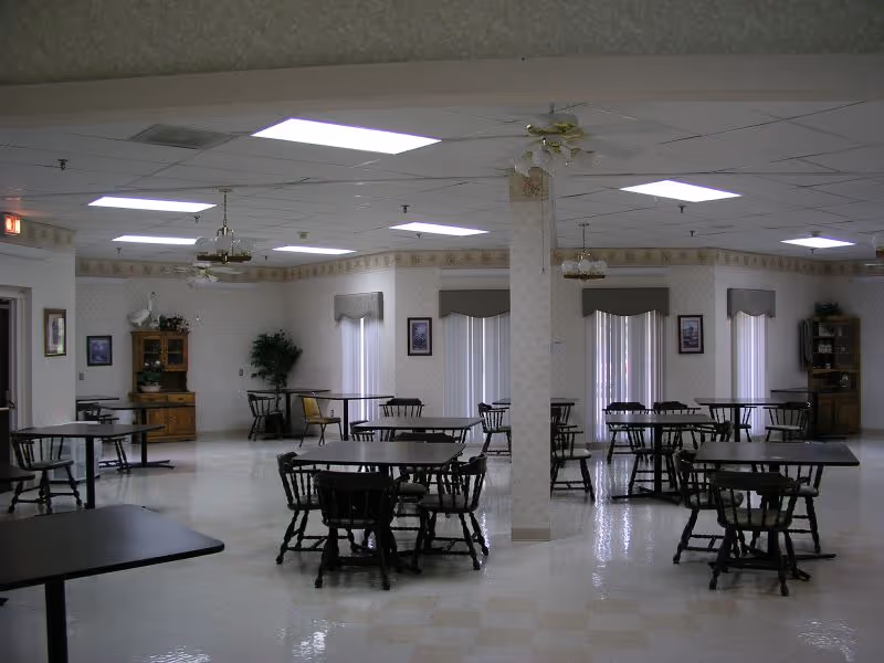 A spacious dining room with multiple dark wooden tables and chairs arranged neatly. The room has a tiled floor, white walls with a floral border near the ceiling, and several windows with vertical blinds and valances. Ceiling fans and fluorescent lights are mounted on the ceiling. There are wooden cabinets and framed pictures on the walls.