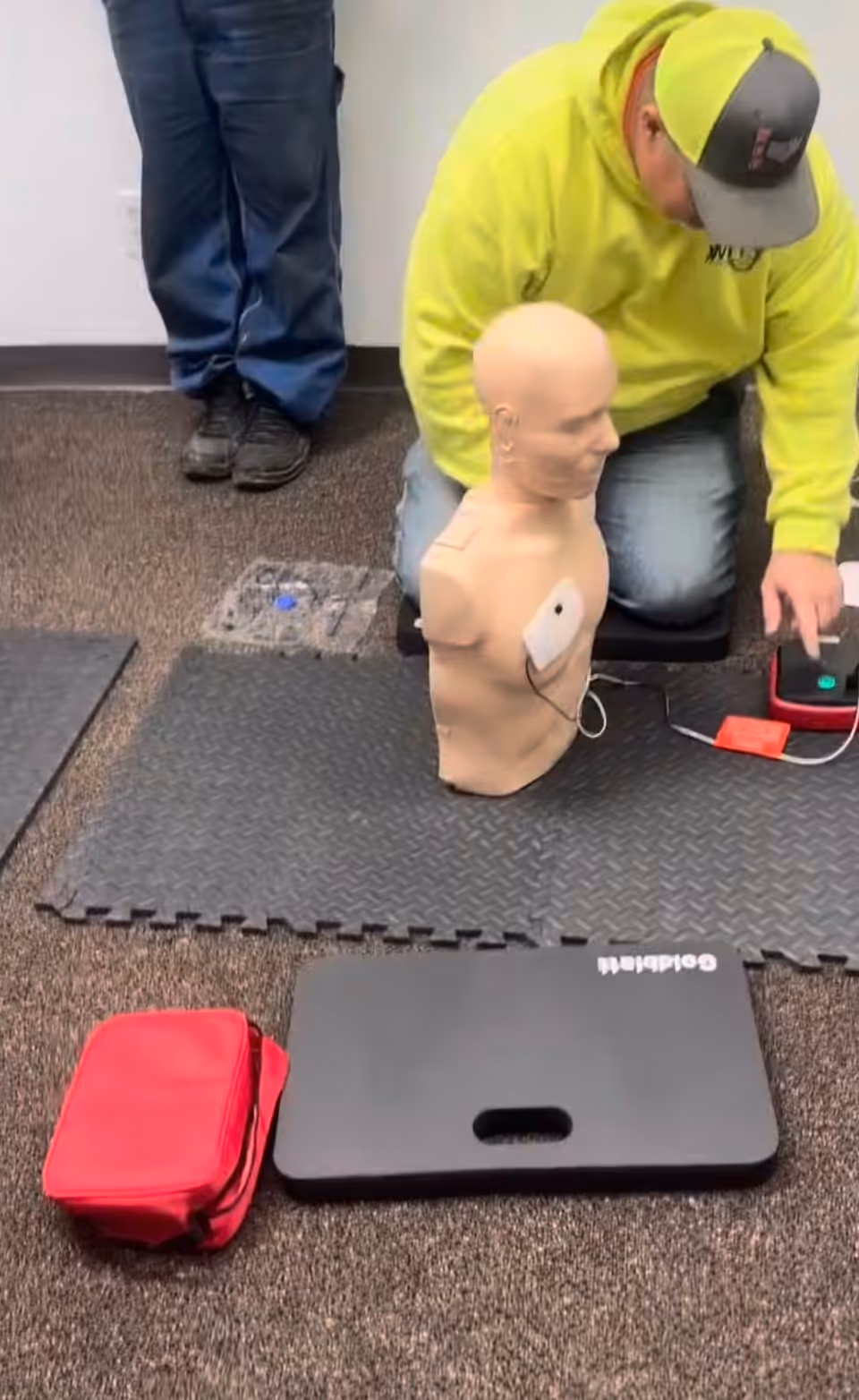 Person in neon hoodie trains CPR on a life-size mannequin with chest pads.