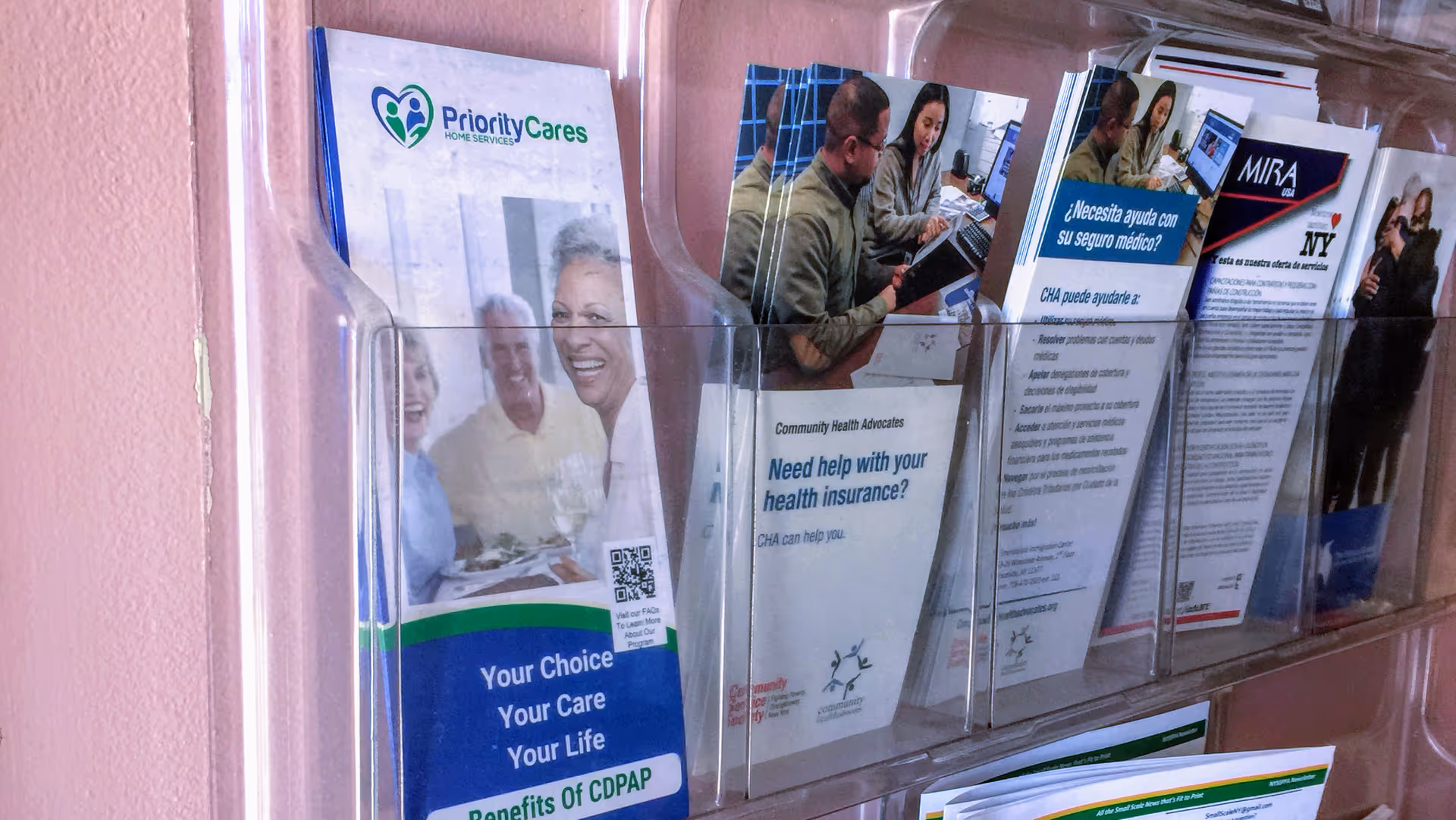 Brochure rack with Priority Cares Home Services pamphlets against a pink wall.
