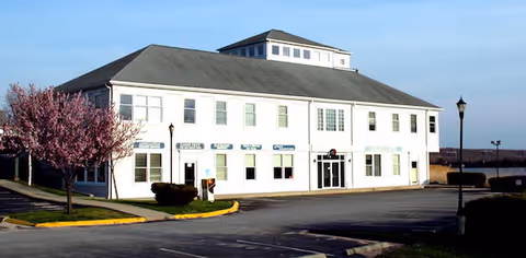 White two-story senior living facility with a blooming tree and parking lot.