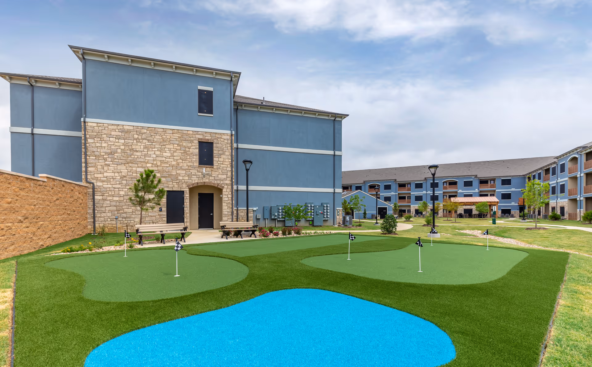 Courtyard of a senior living facility featuring a small putting green with flags, a blue decorative pond, benches, and surrounding multi-story blue buildings.