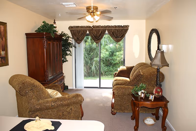 A cozy living room with two upholstered armchairs and a matching sofa arranged around a wooden side table with a lamp and framed photos. There is a large wooden cabinet on the left side and a sliding glass door with brown curtains that opens to a green outdoor area. A ceiling fan with lights is mounted on the ceiling.
