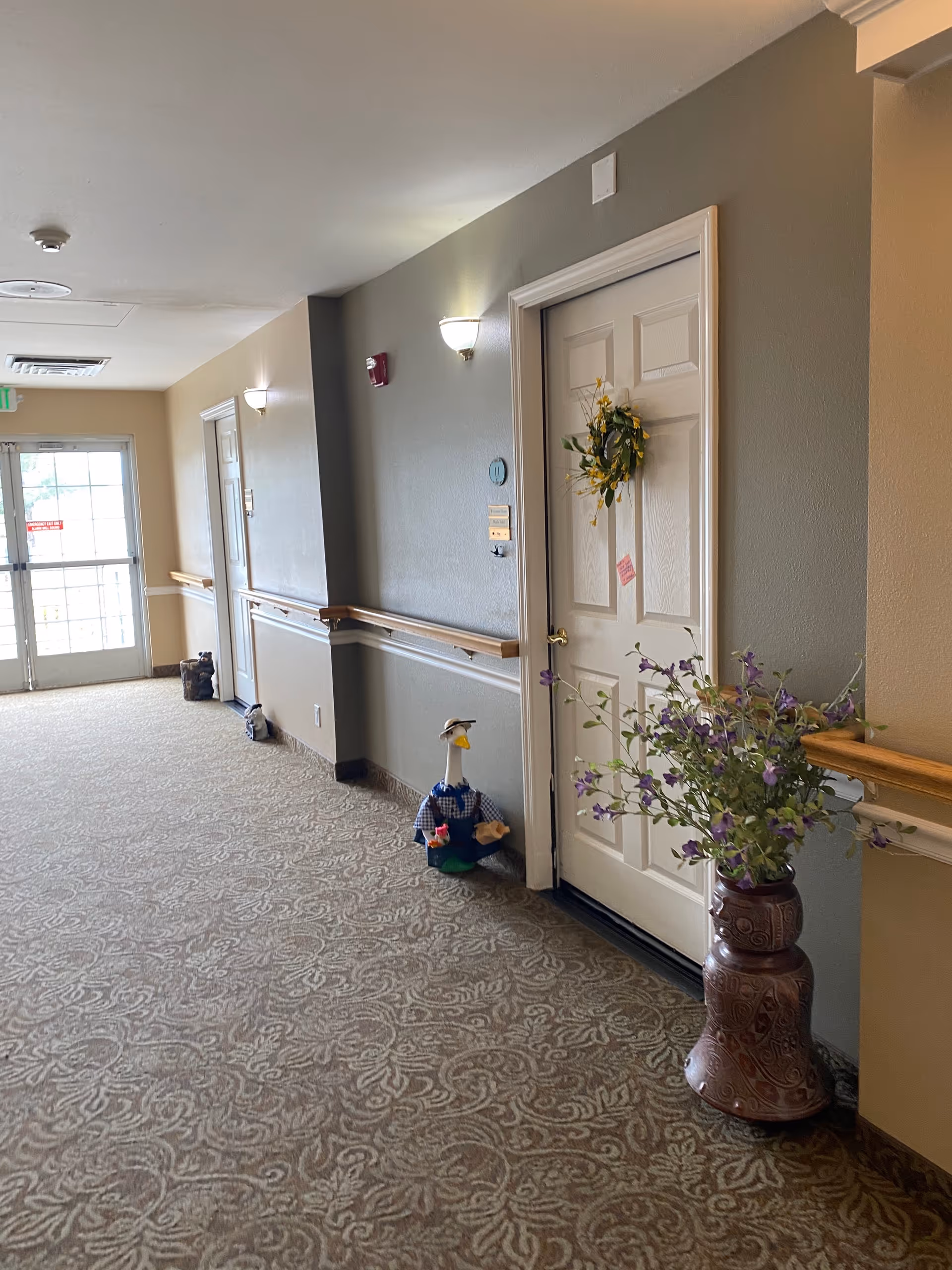 Interior hallway of Volante Senior Living with wreath on door, vase of flowers, and patterned carpet.