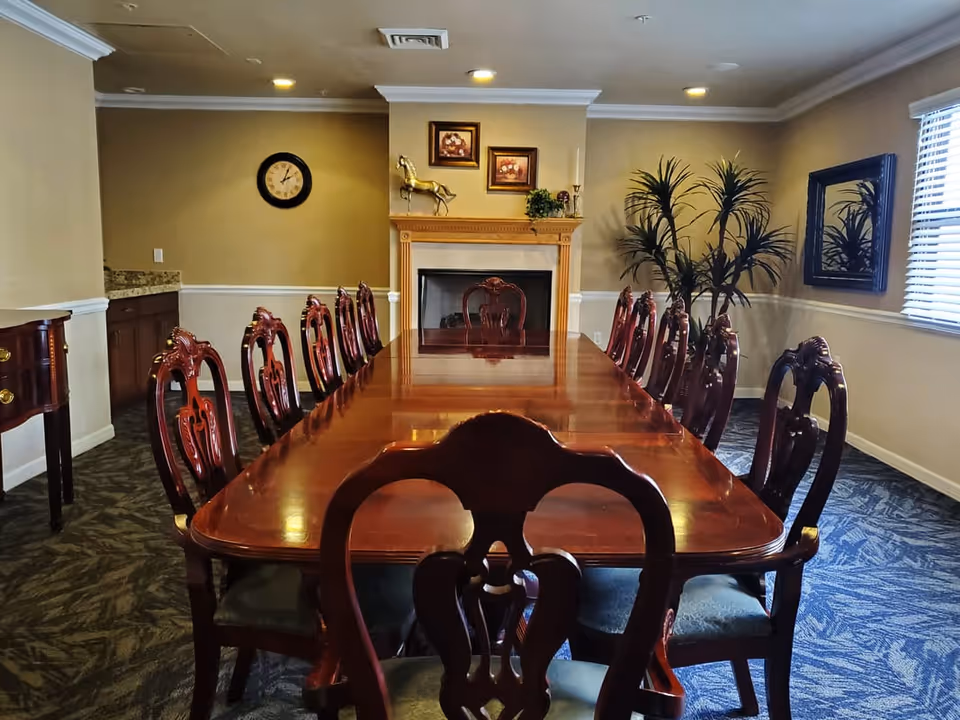 A formal dining room with a long polished wooden table surrounded by twelve ornate wooden chairs. The room features a fireplace with decorative items and framed pictures above it, a wall clock, a large mirror, and a potted plant near a window with blinds. The carpet has a patterned design and the walls are painted beige with white trim.