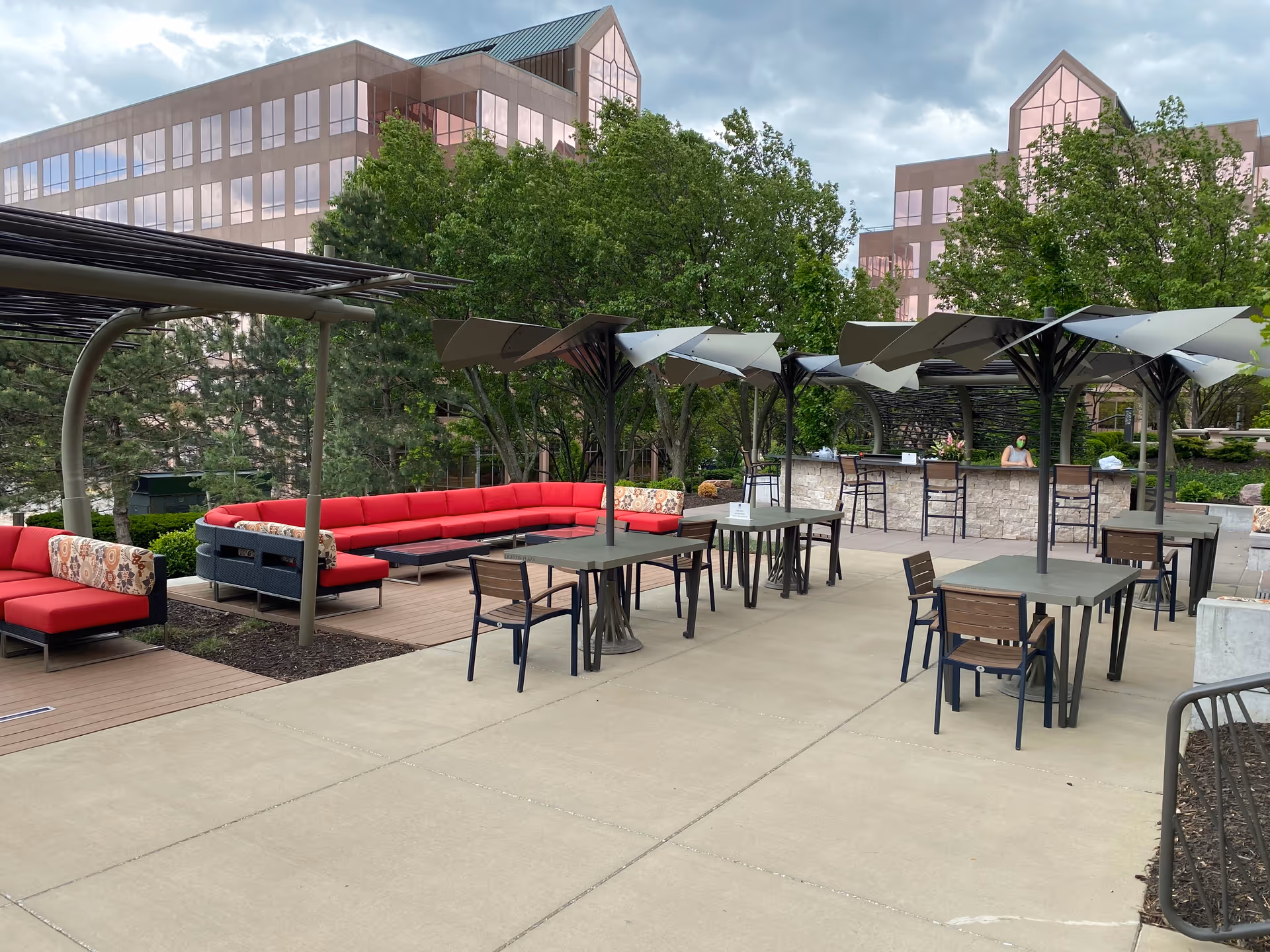 Outdoor patio with umbrellas, tables, chairs, and red cushioned seating beside a modern building.