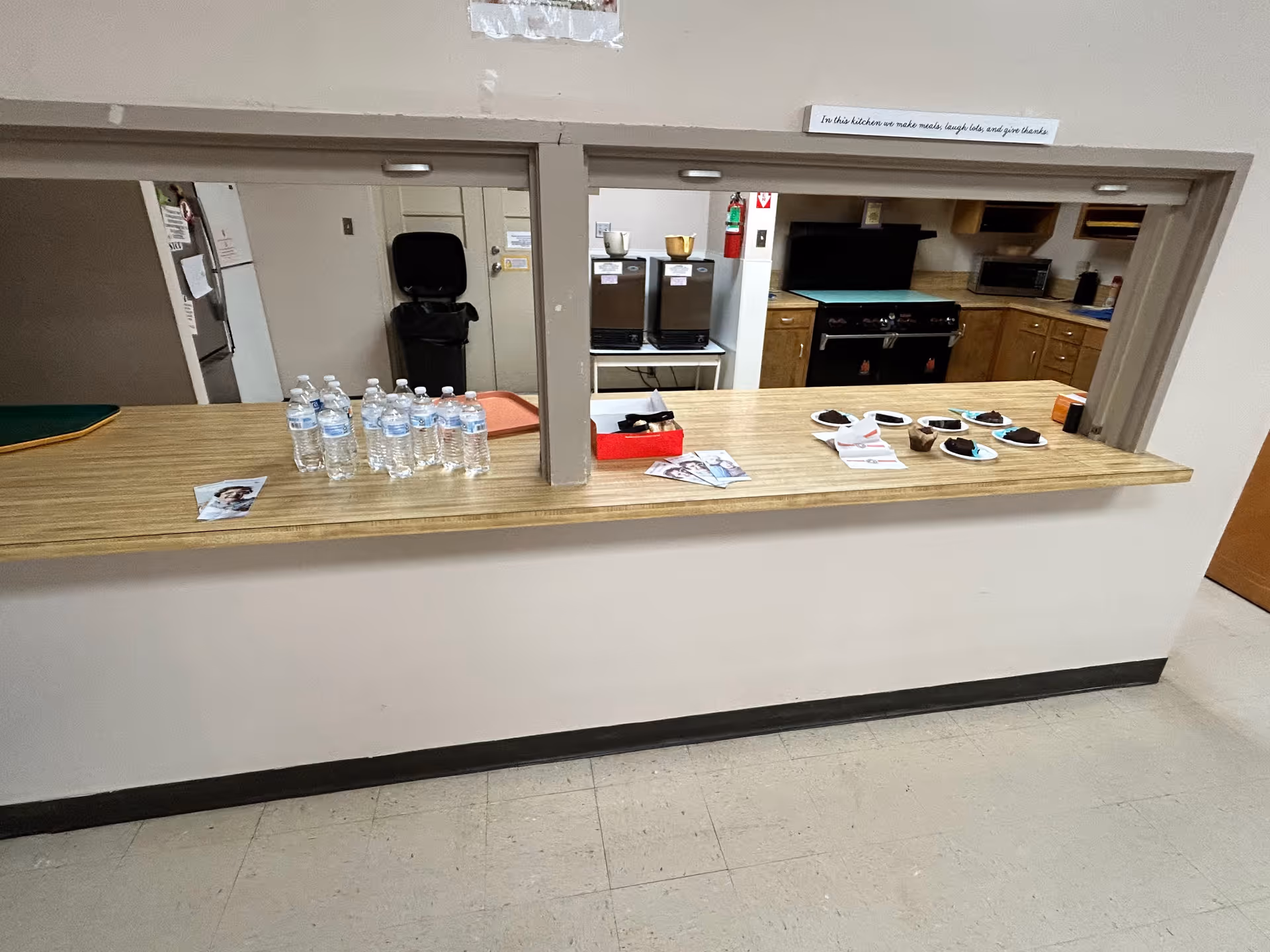 Service counter with bottled water and desserts, kitchen appliances visible in senior living facility.