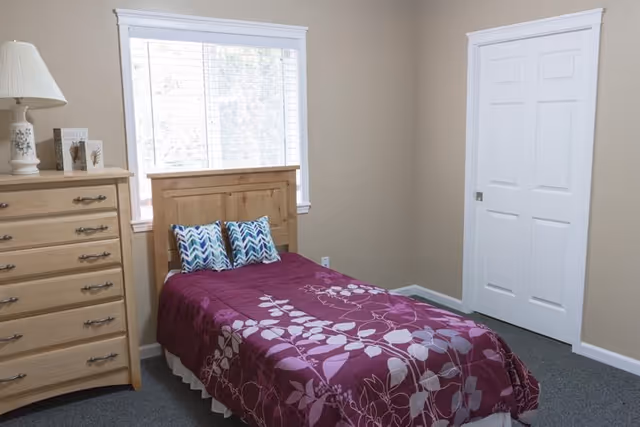 A small bedroom with a single wooden bed covered with a purple floral bedspread and two patterned pillows. Next to the bed is a wooden dresser with a white lamp and decorative items on top. A large window with white blinds is behind the bed, and a white closet door is on the right wall.