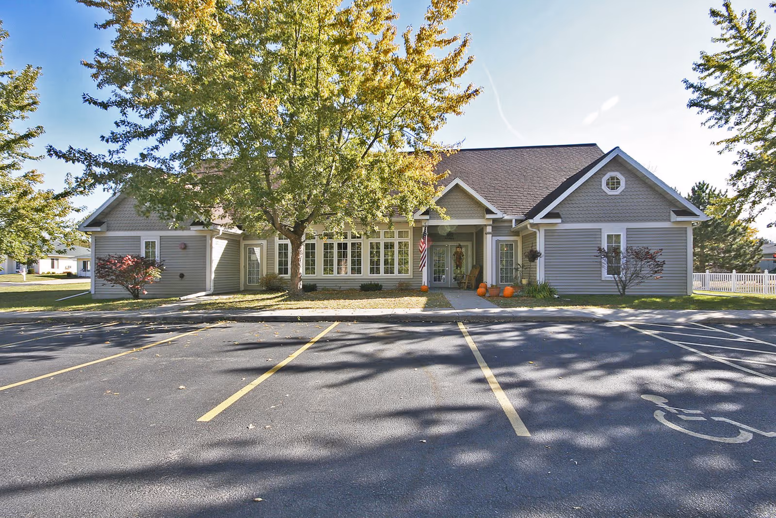 Front exterior view of a single-story senior living facility building with gray siding, white trim, and a dark shingled roof. There is a large tree with green leaves in front of the building, a sidewalk leading to the entrance, and several pumpkins near the door. The parking lot in front has marked spaces including a handicapped spot.