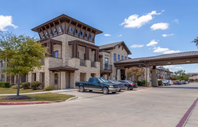 Stone senior living building with a covered entrance and parked cars under a blue sky.