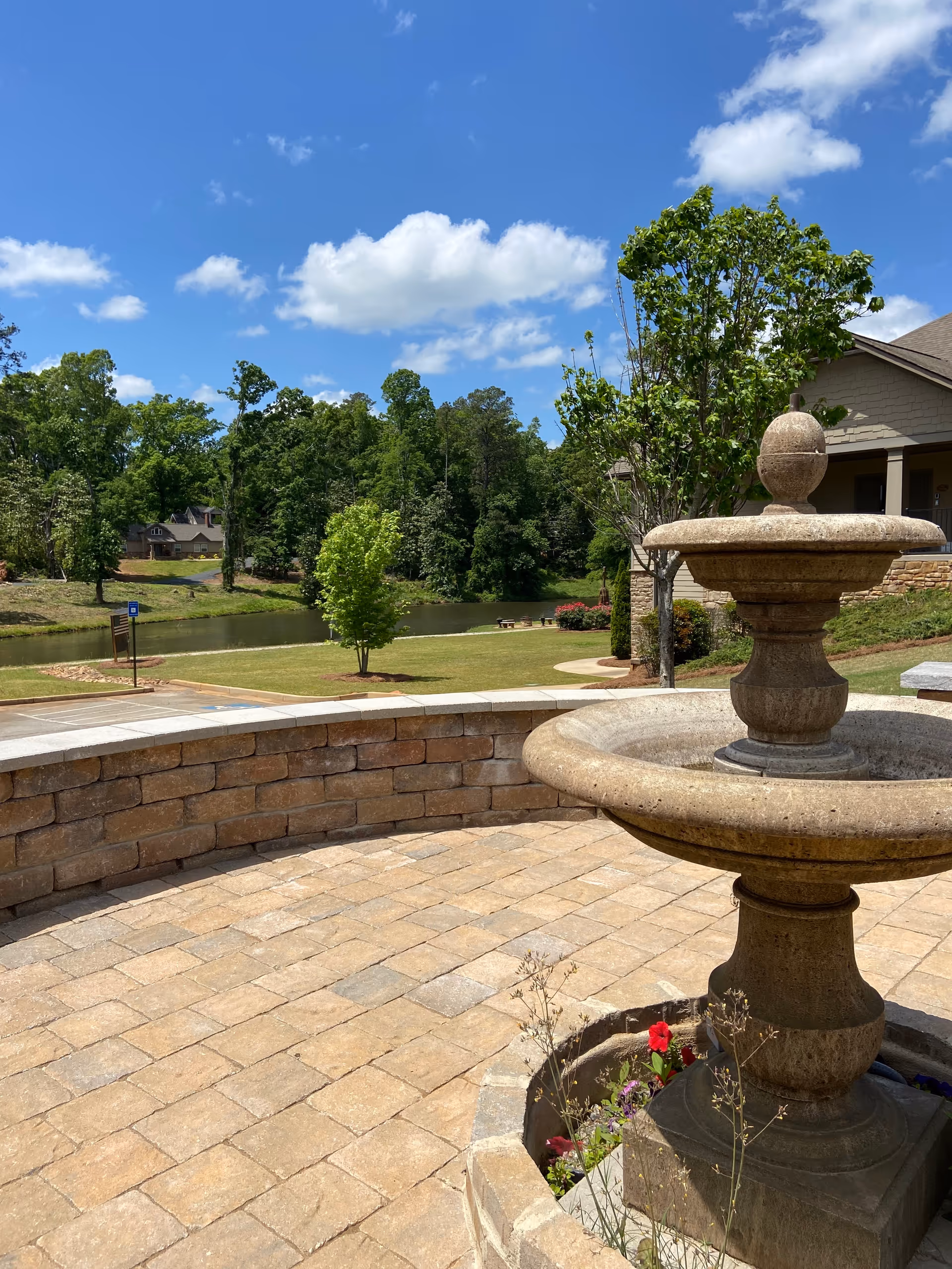 Sunlit stone courtyard with a tiered fountain, brick edging, and a scenic pond.
