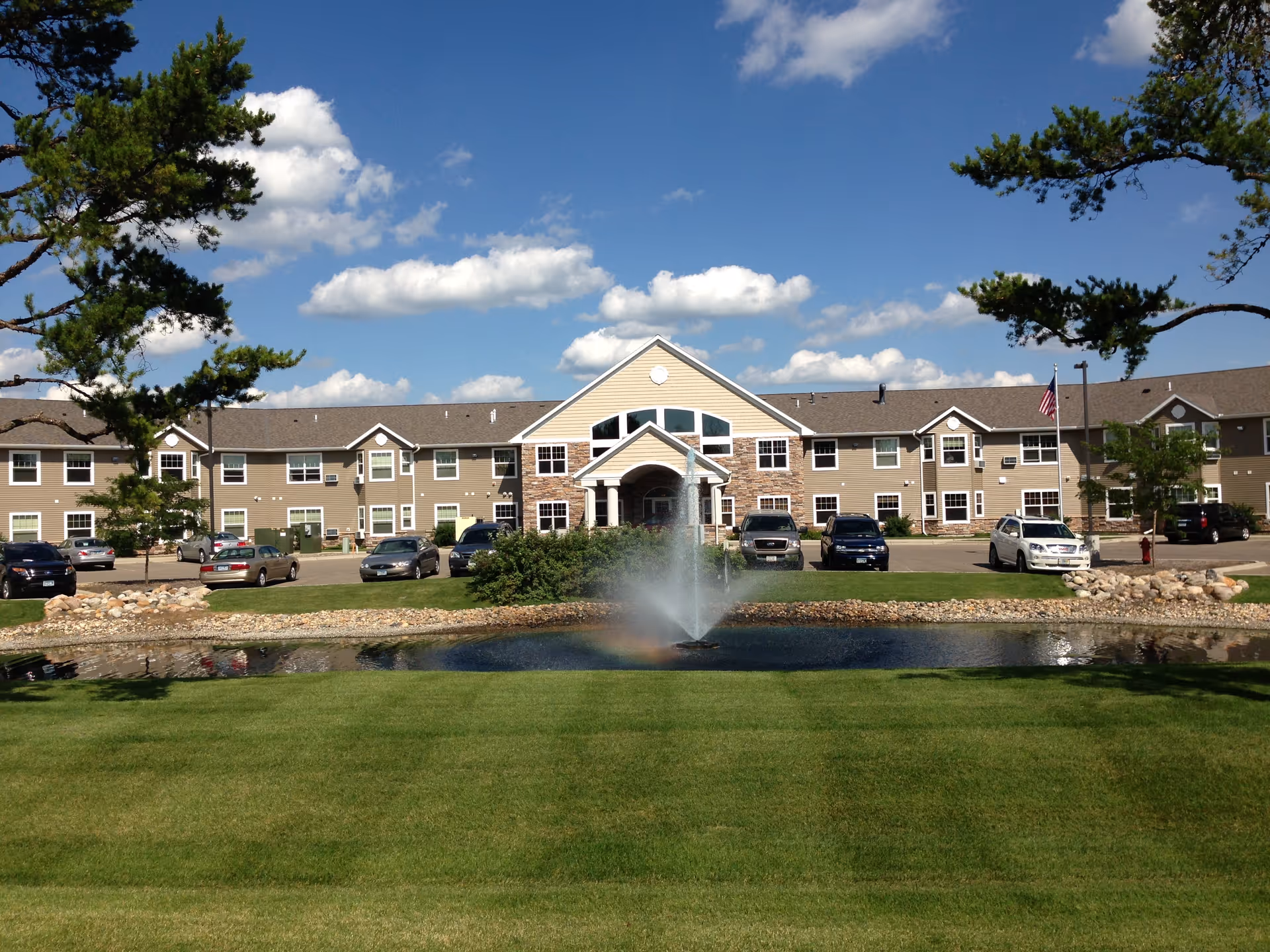 Senior living facility building with a fountain pond, parked cars, and landscaped lawns.