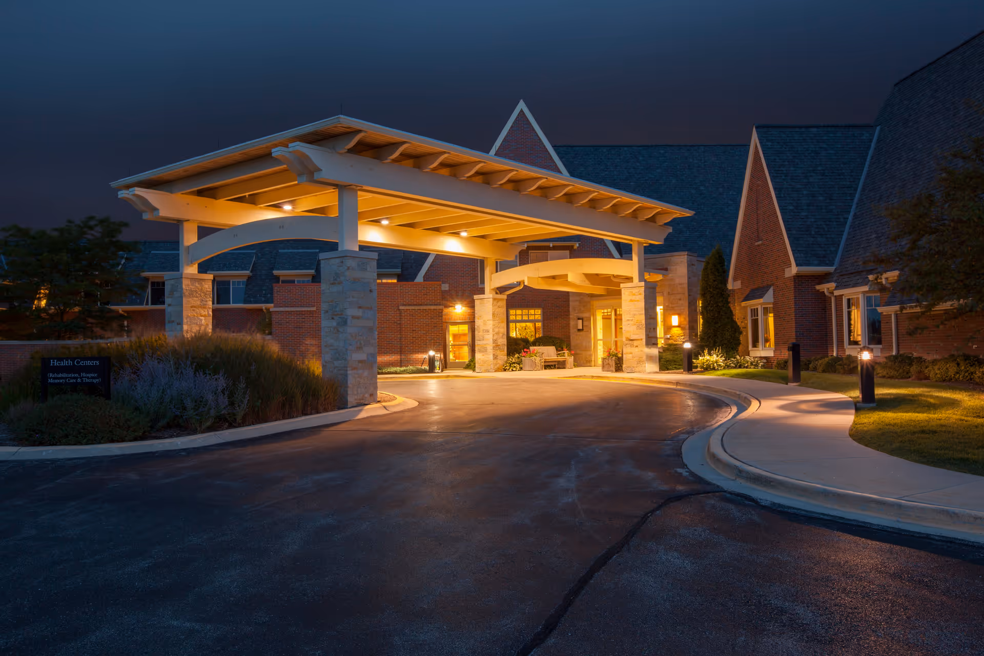 Entrance of a senior living facility named Teal Shores at dusk, featuring a covered drop-off area with stone pillars and warm lighting. The building has brick walls and steeply pitched roofs, with a curved driveway and landscaped greenery around the entrance.