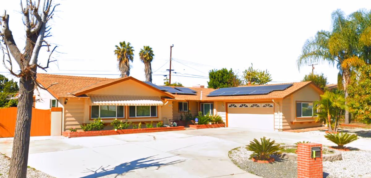Single-story residential building with a light brown exterior, solar panels on the roof, a two-car garage, and a driveway surrounded by desert landscaping with palm trees and small shrubs.