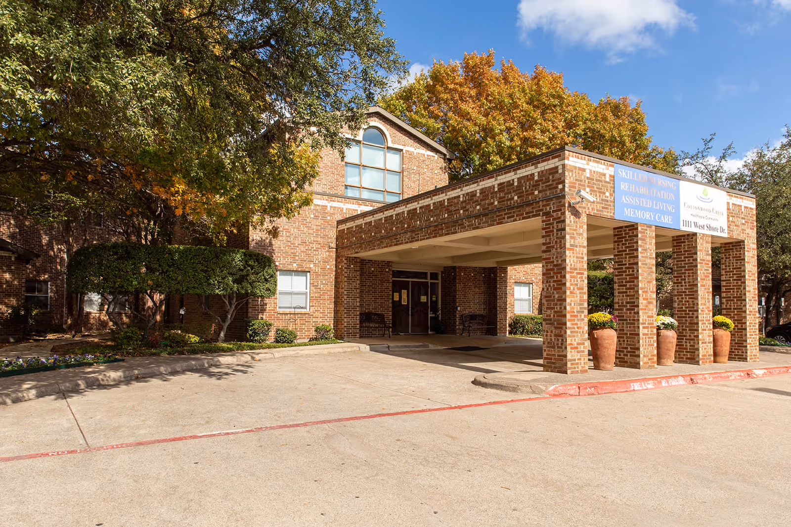 Exterior view of Cottonwood Creek Healthcare Community showing a brick building entrance with a covered drop-off area. There are large windows, trimmed bushes, potted plants with flowers, and trees with autumn foliage under a clear blue sky.