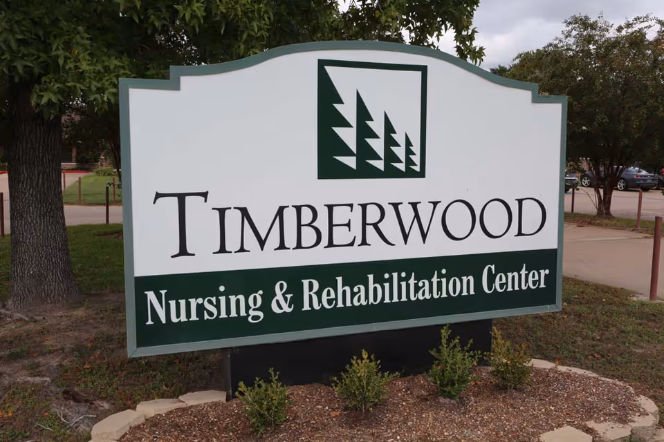 A large outdoor sign for Timberwood Nursing & Rehabilitation Center with a logo featuring stylized pine trees. The sign is surrounded by small plants and trees, with a parking area and sidewalk visible in the background.