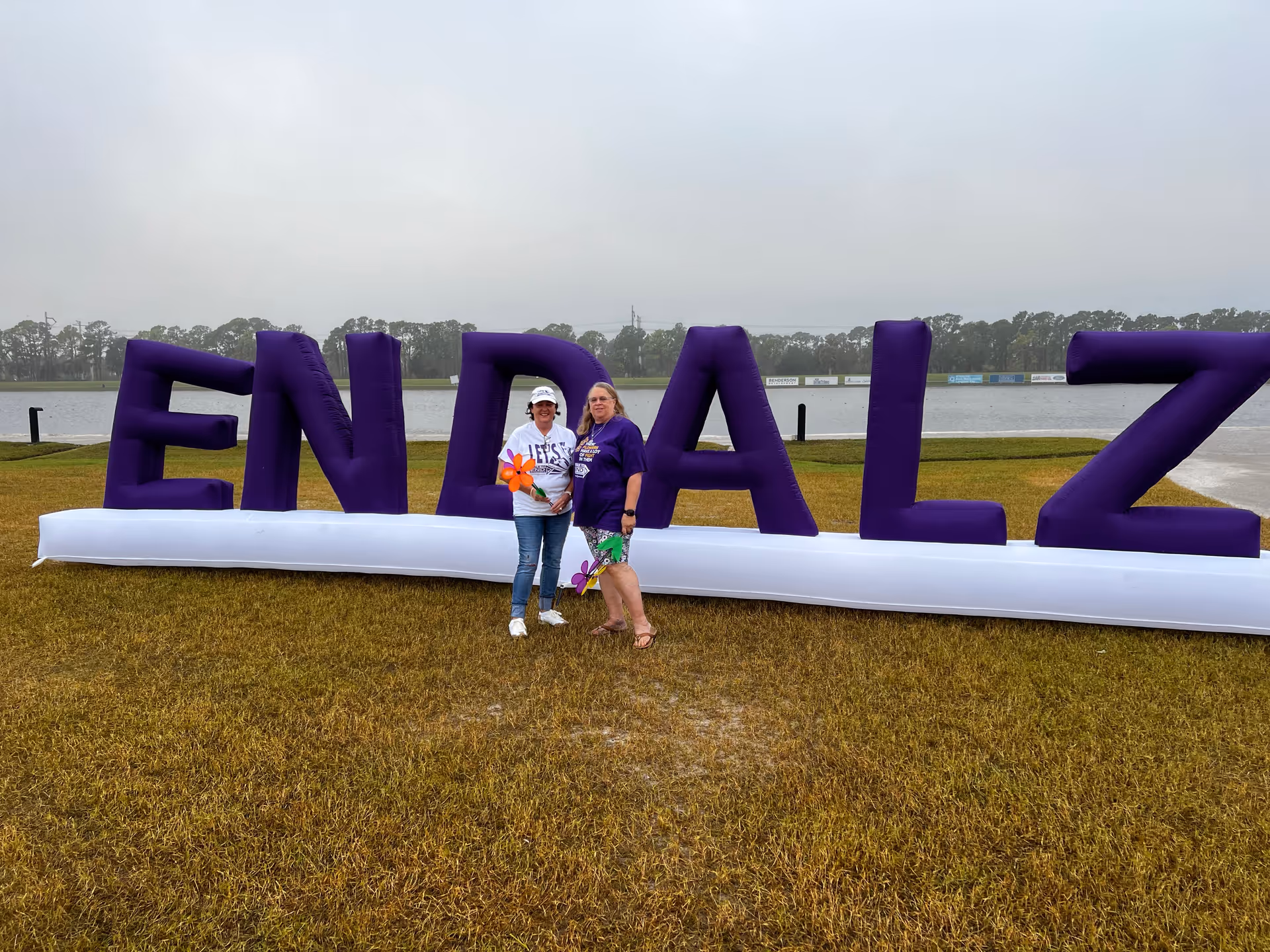 Two people pose beside large purple inflatable letters spelling ANGELS near a lake.