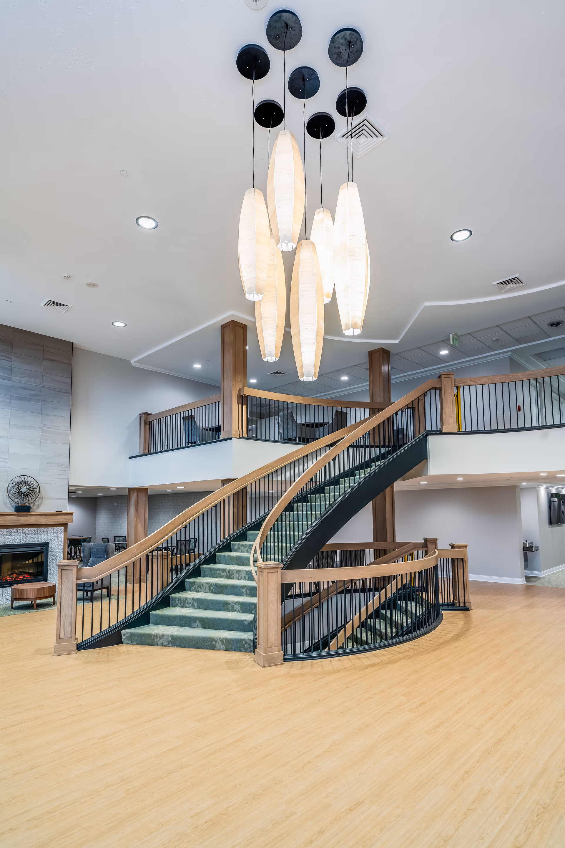 Interior view of a senior living facility featuring a wide staircase with green carpet and wooden handrails leading to an upper level. Above the staircase hangs a modern chandelier with multiple elongated white pendant lights. The space has light wood flooring, a fireplace with seating area to the left, and a high ceiling with recessed lighting.