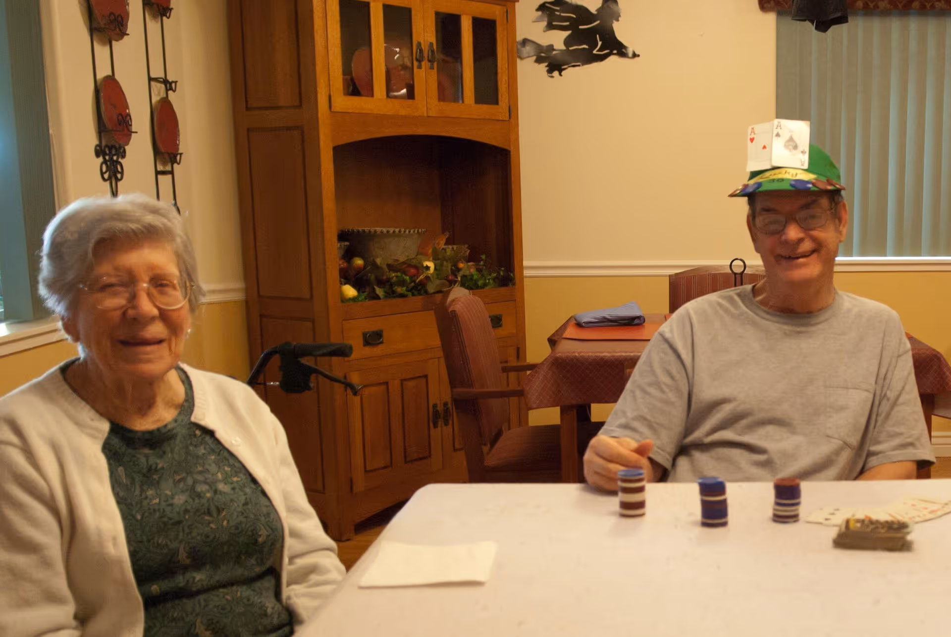 Two elderly individuals sitting at a table in a room. The woman on the left is wearing glasses, a green patterned top, and a white cardigan. The man on the right is wearing glasses, a gray t-shirt, and a colorful hat with playing cards attached to it. There are poker chips and playing cards on the table in front of them. Behind them is a wooden cabinet with decorative items and a wall with some artwork and a window with vertical blinds.