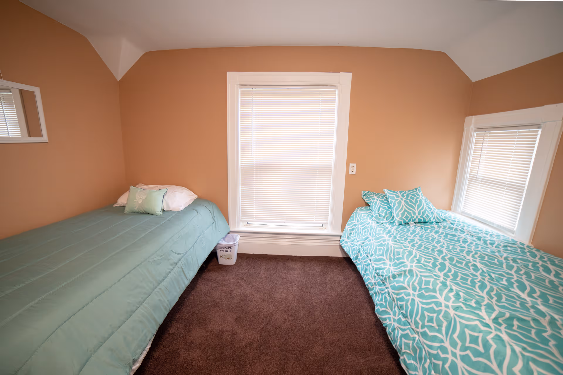 Two twin beds with teal bedding flank windows in a beige bedroom.