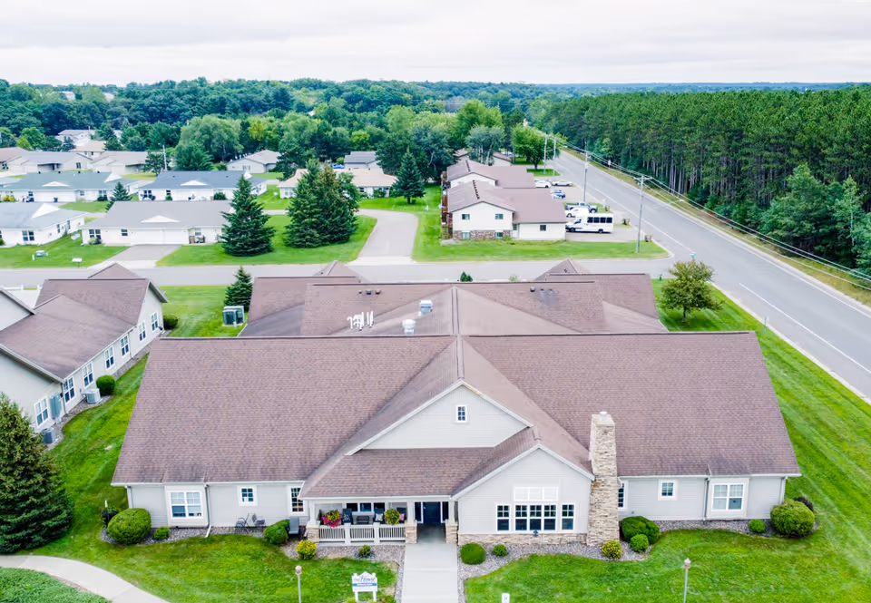 Aerial view of a single-story senior living facility building with a brown roof and light-colored exterior walls, surrounded by green lawns, trees, and a paved road on the right side. Additional residential buildings and a wooded area are visible in the background under a cloudy sky.