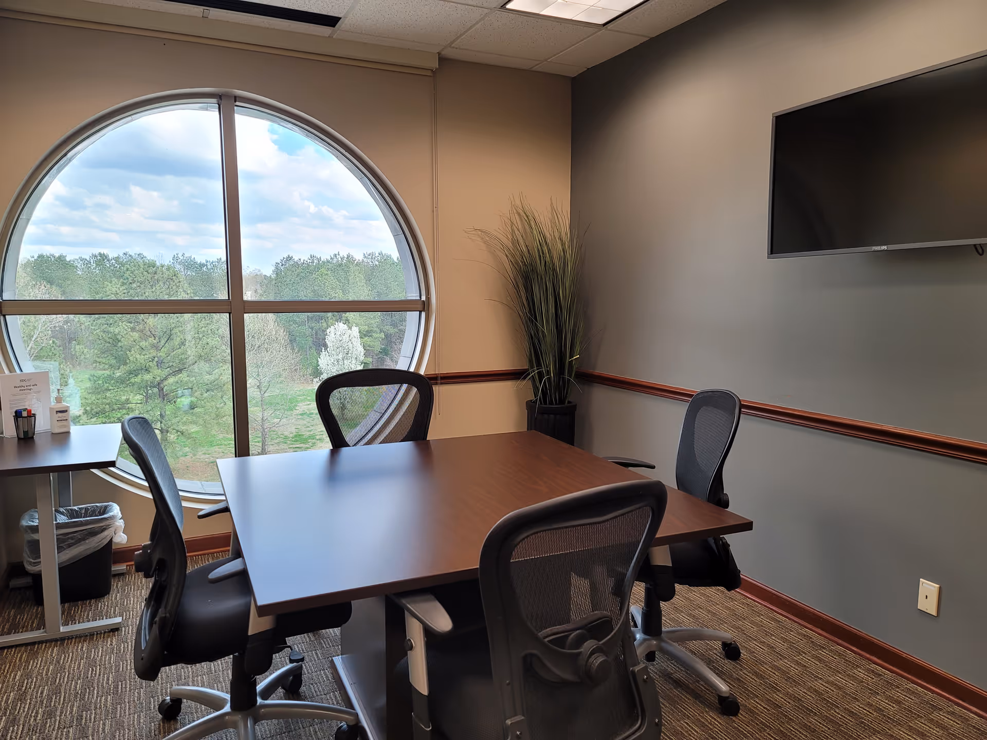 Office-like meeting room with round window, table, chairs, and wall-mounted TV.