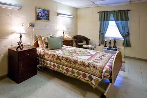 A furnished patient bedroom in a nursing home with a quilted bed, nightstands, and a window.