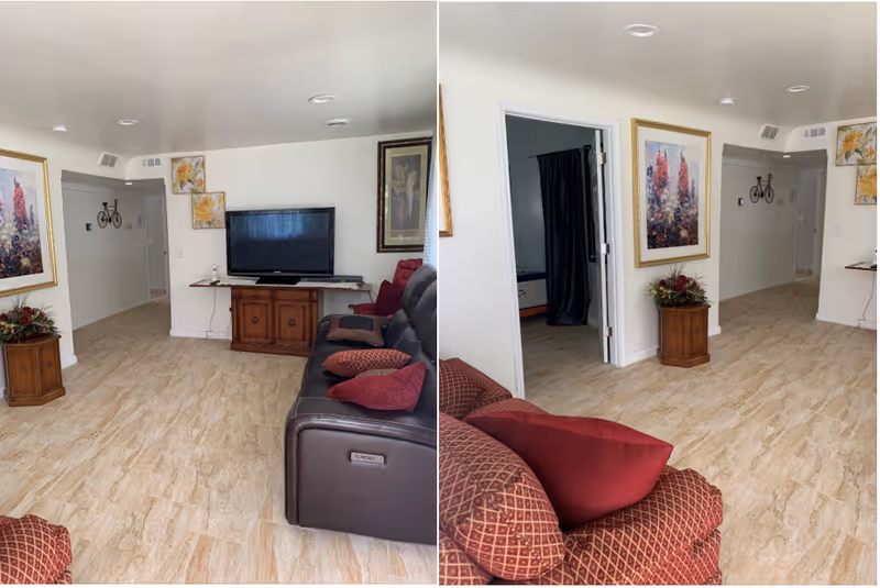 Two views of a living room area with light wood flooring, white walls, and recessed ceiling lights. The room features a dark brown leather sofa with red and patterned cushions, a wooden TV stand with a flat-screen TV, and framed floral artwork on the walls. A small wooden side table with a floral arrangement is positioned near the entrance to a hallway and an open doorway with a dark curtain.