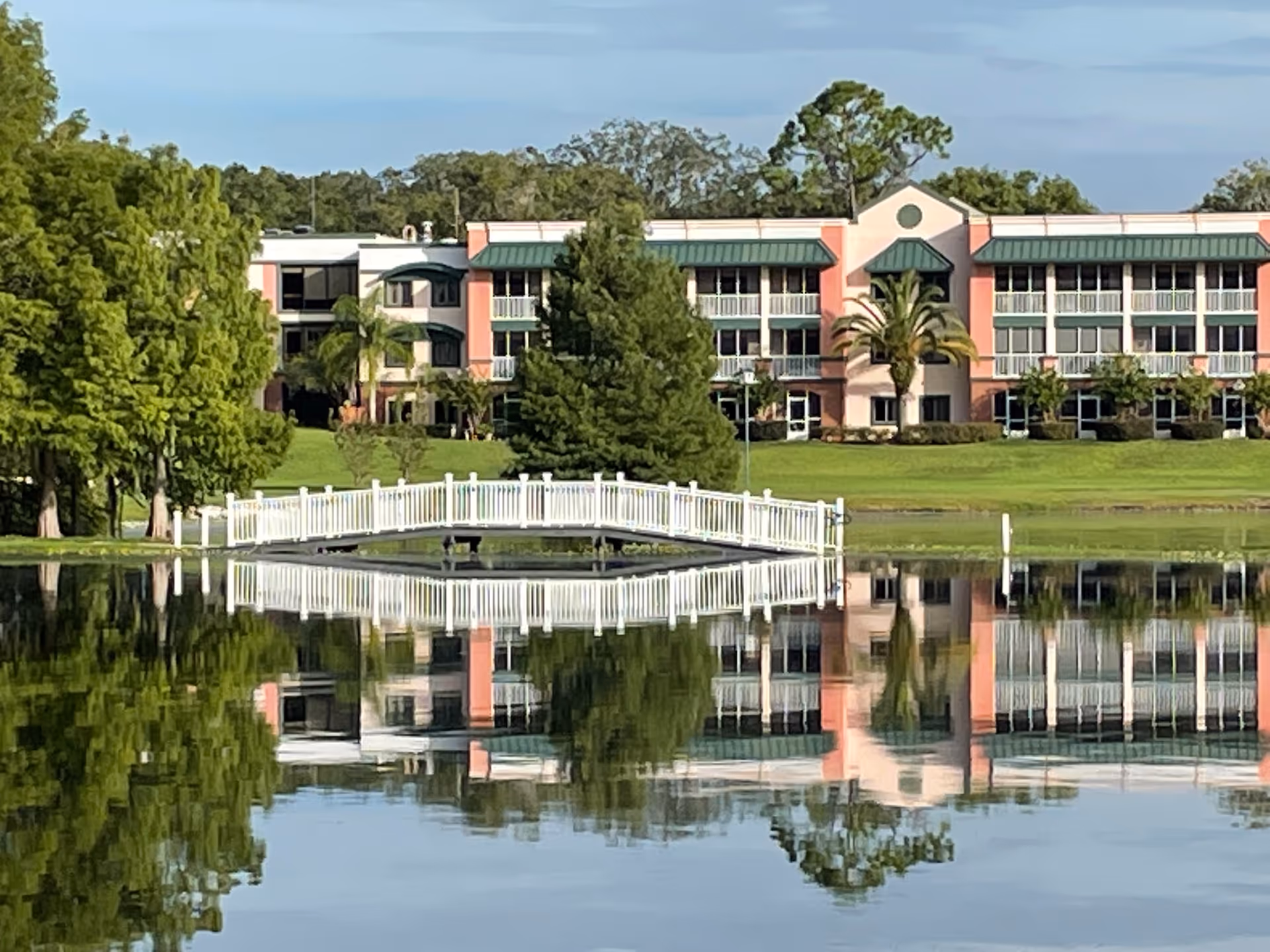 Senior living campus building with pink facade, palm trees, and a white footbridge over water.