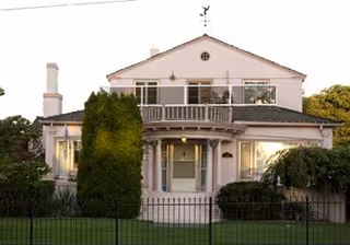 Two-story house with a front porch, balcony, and surrounding shrubs behind a black fence.