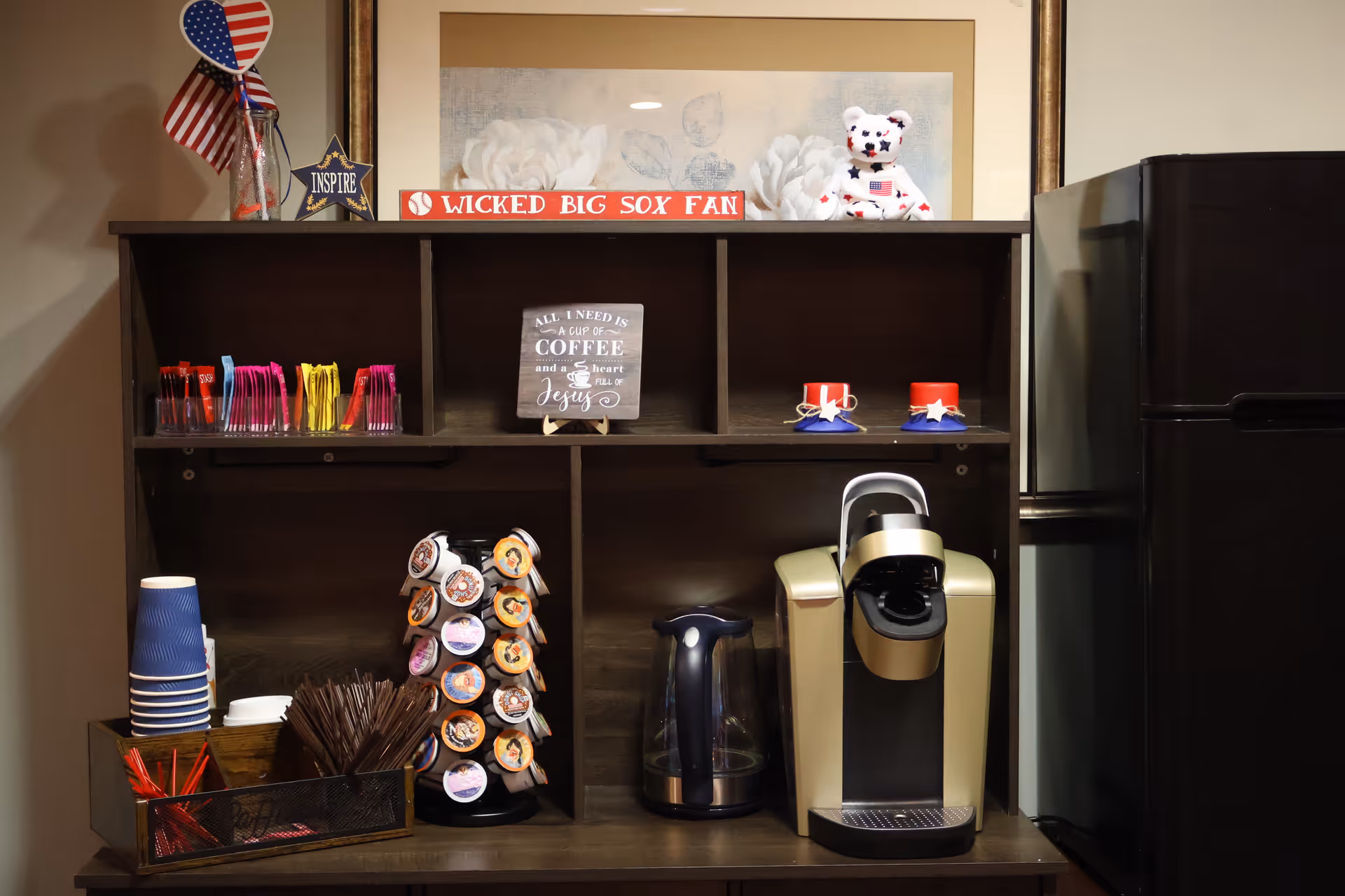 A coffee station with a coffee maker, electric kettle, and a rotating holder filled with coffee pods. There are disposable cups, stirrers, and packets of sugar and creamer. On the top shelf, there are patriotic decorations including small American flags, a star with the word 'INSPIRE', a sign that reads 'WICKED BIG SOX FAN', and a small teddy bear with an American flag design. A framed floral artwork hangs on the wall behind the station.