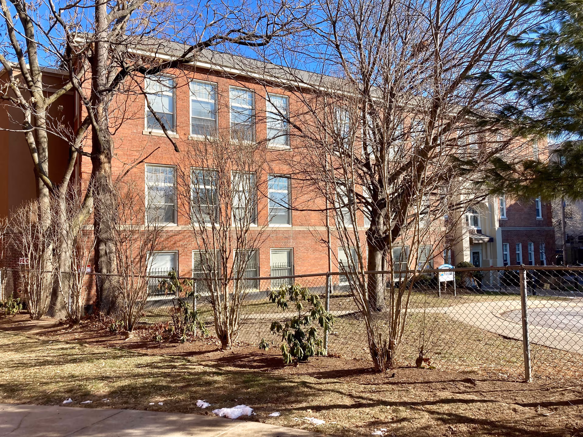 Front exterior of a three-story red brick assisted living building behind a chain-link fence with leafless trees and a small lawn.