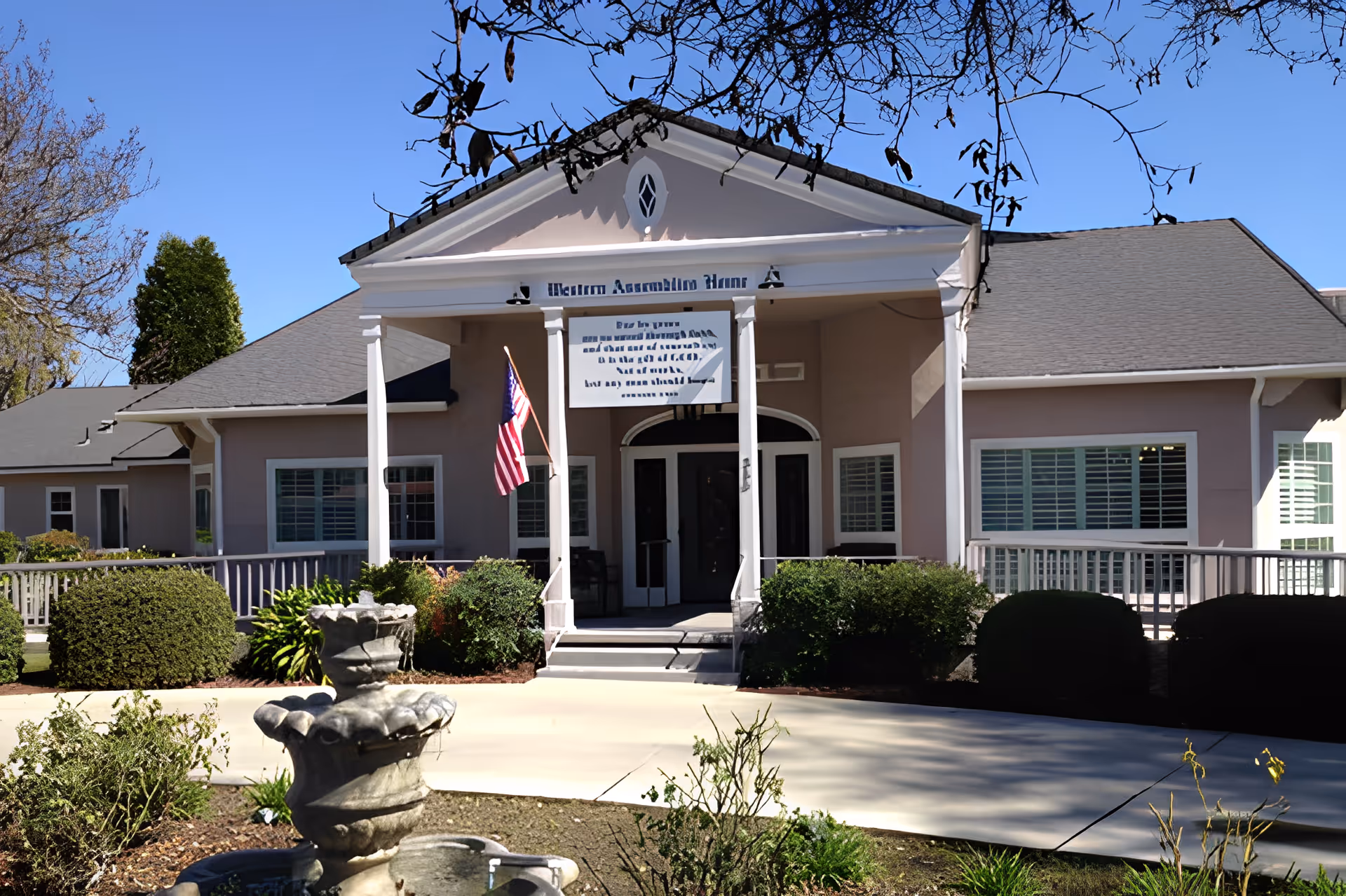 Front entrance of a single-story senior living facility with columns, an American flag, shrubs, and a fountain in the foreground.