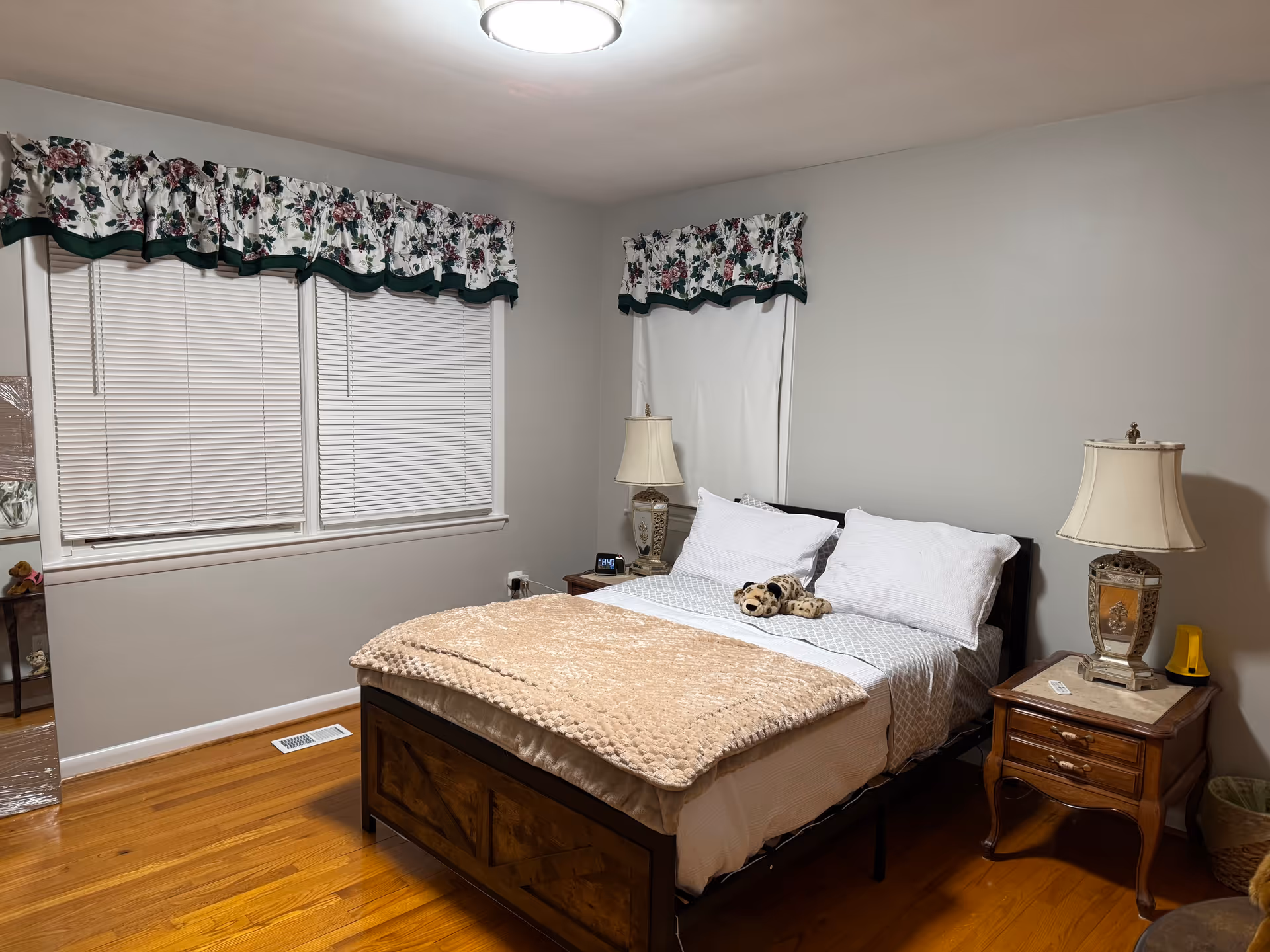 A neatly made bedroom with a bed, nightstands, lamps, and floral window valances.