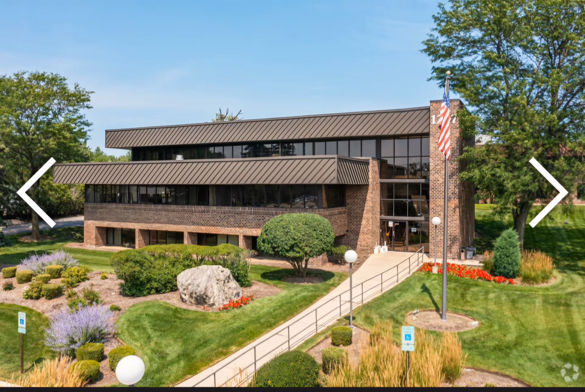 Modern brick senior living building with manicured grounds, flagpole, and large windows.