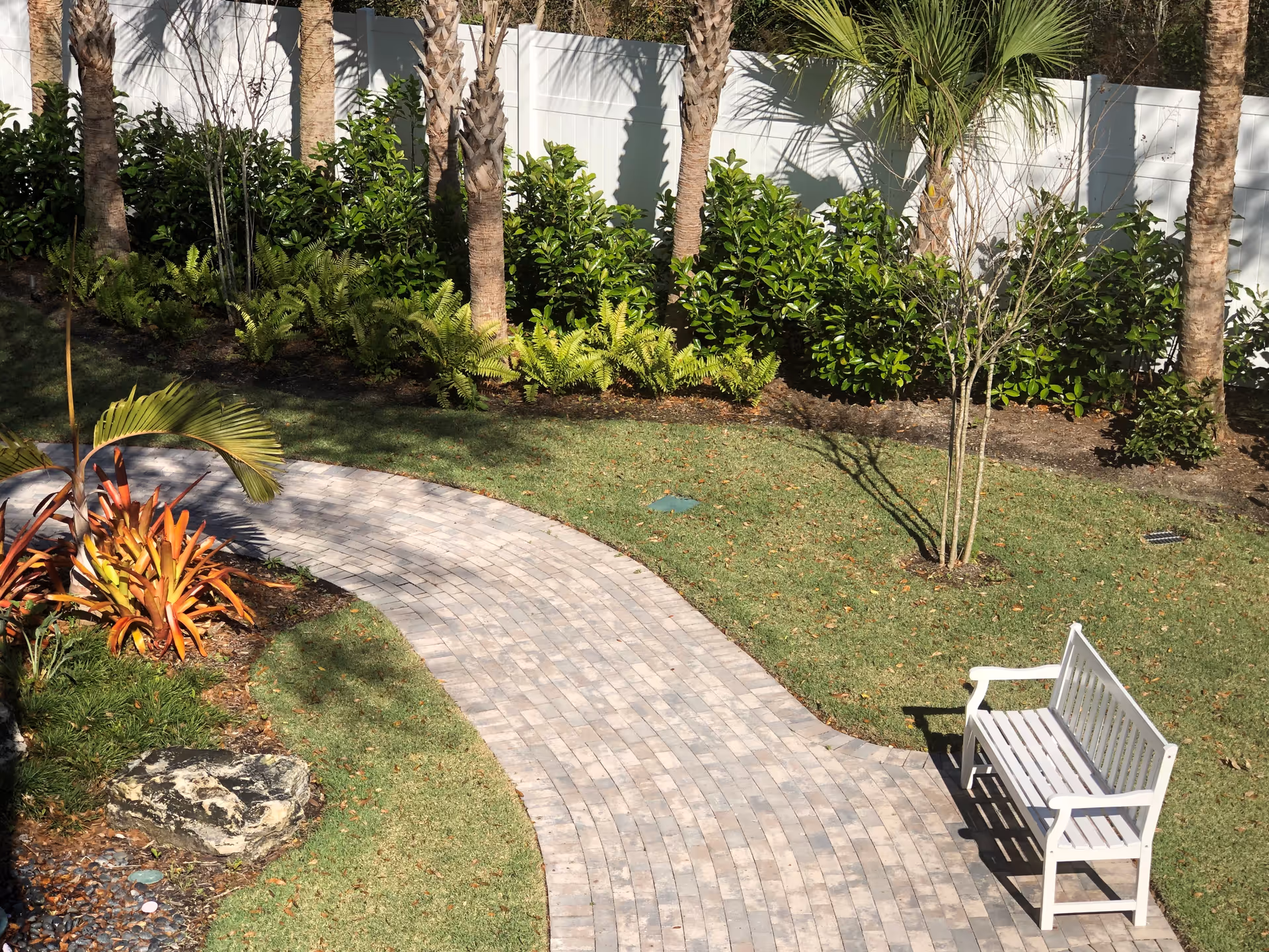 A curved paved walkway in a garden area with green grass, tropical plants, palm trees, and a white wooden bench on the right side. A white fence is visible in the background.
