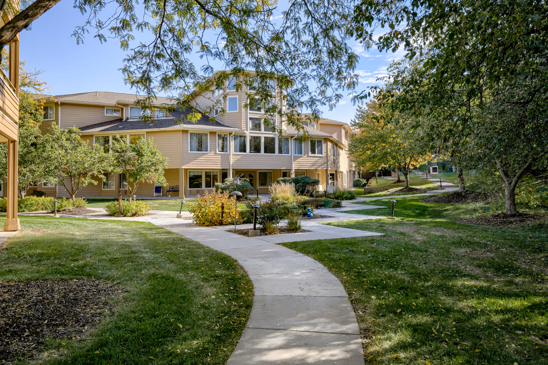 A landscaped courtyard with a paved walkway leading to a multi-story beige senior living building surrounded by trees and lawns.