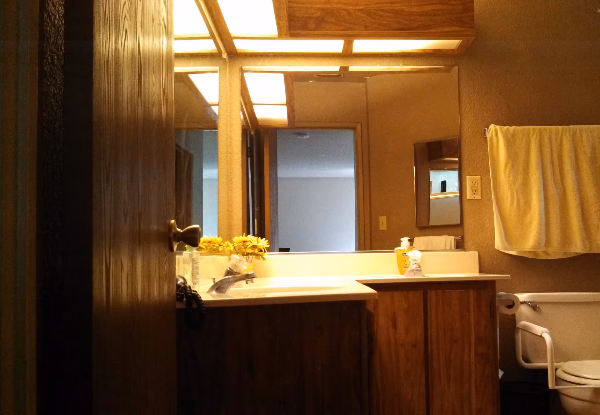 Interior view of a bathroom with a wooden door partially open. The bathroom features a sink with a countertop, a mirror above the sink, a towel hanging on a rack, a toilet with a safety grab bar, and some yellow flowers on the counter. The lighting is warm and the walls are textured.