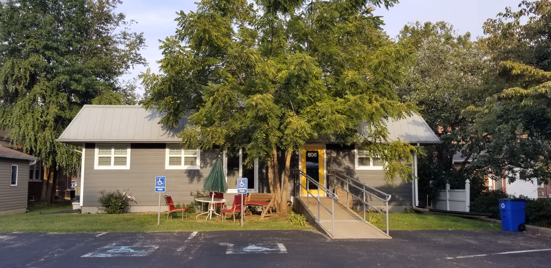 Single-story assisted living facility with ramp, wheelchair-accessible parking, and outdoor seating under trees.