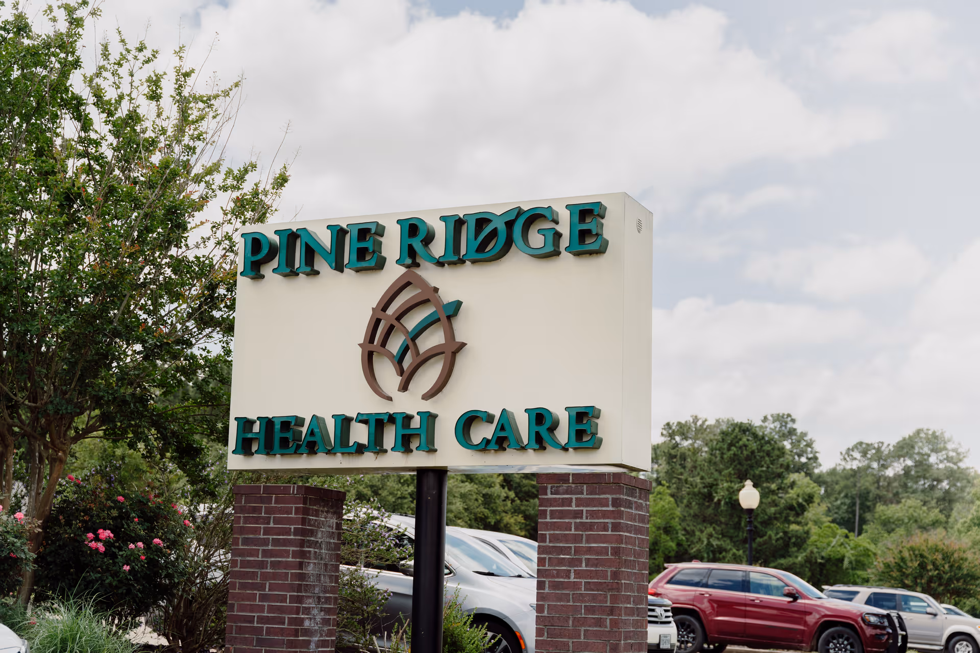 Outdoor view of a sign for Pine Ridge Health Care mounted on two brick pillars with trees, parked cars, and a cloudy sky in the background.