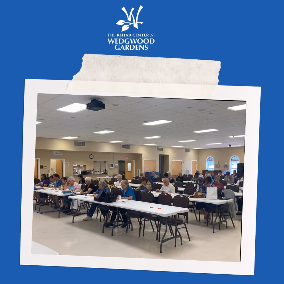 A large room with many elderly people seated at long tables, engaged in activities or writing. The room has a high ceiling with fluorescent lights, beige walls, and several doors and windows. The setting appears to be a community or activity room within a senior living or rehab facility.
