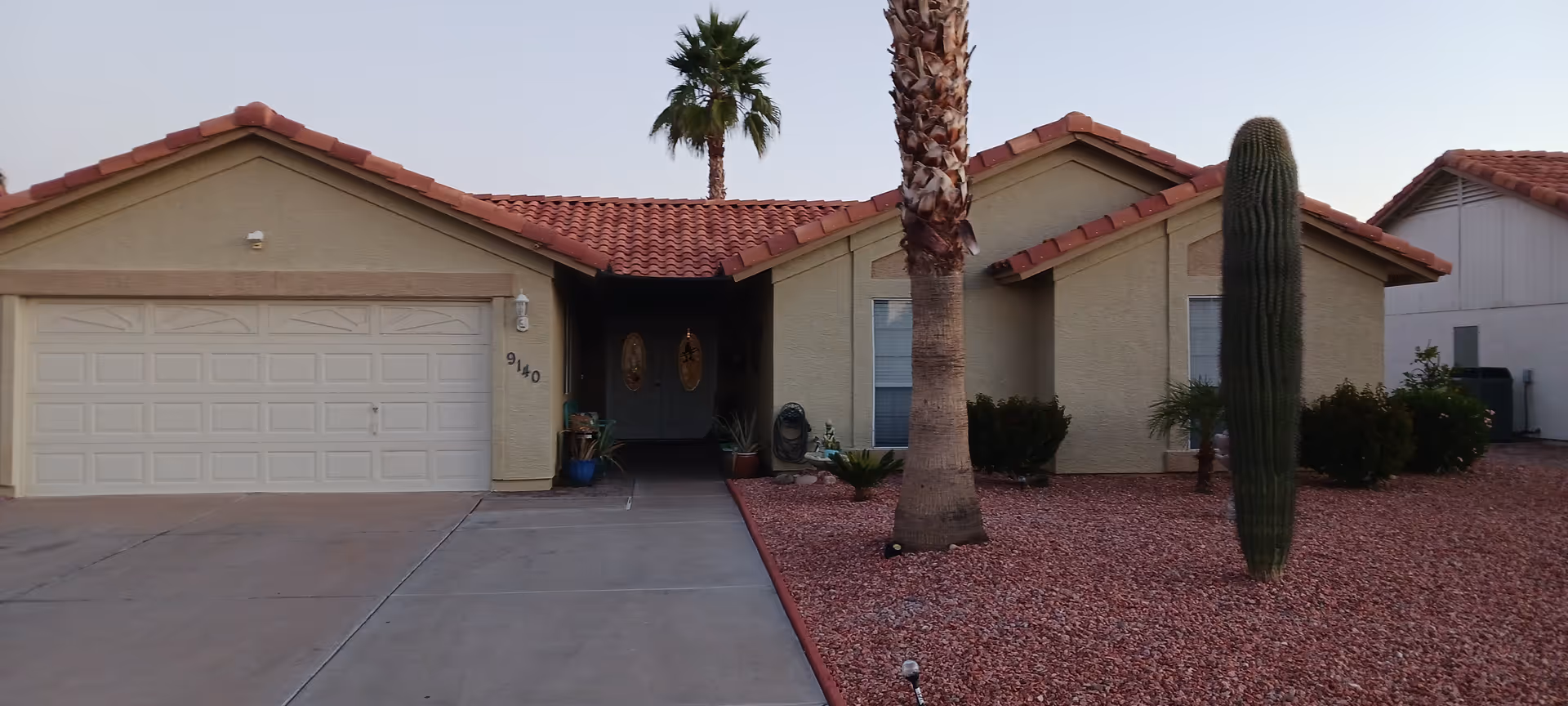 Single-story beige building with red-tiled roof, driveway, garage, palm tree, and desert landscaping.