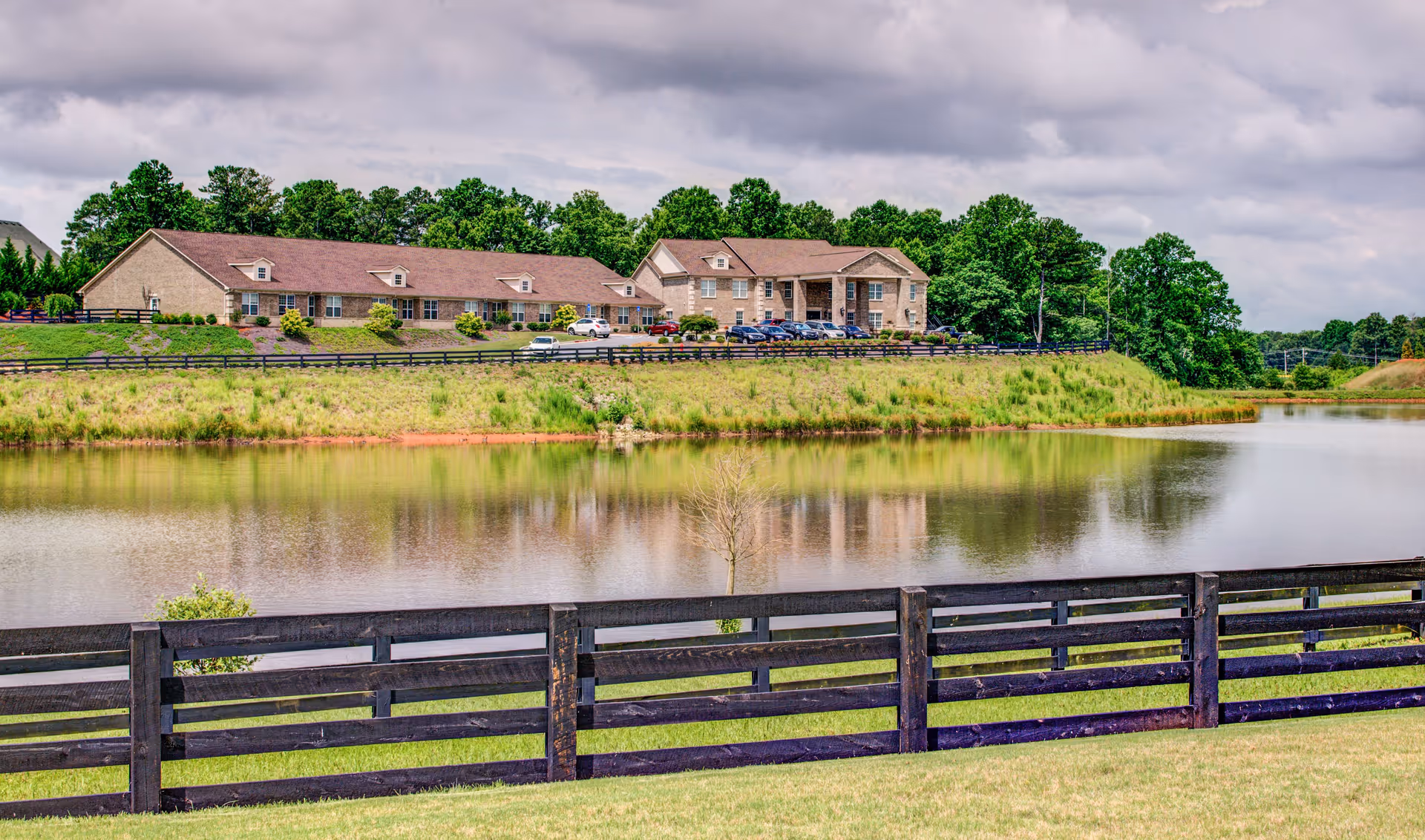 A large senior living facility sits along a pond with a wooden fence in the foreground.
