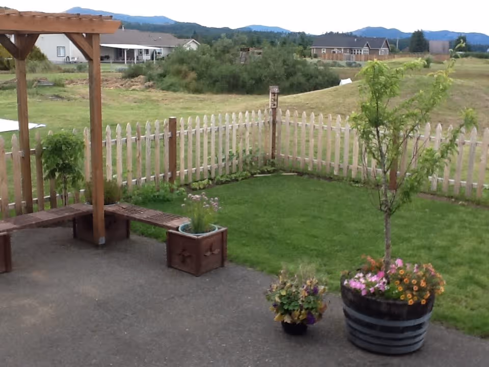 Outdoor patio with wooden pergola, bench, potted flowers, and a white picket fence surrounding a lawn.