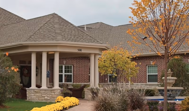 Front entrance of a brick senior living building with a covered portico, columns, landscaping, and a fountain.