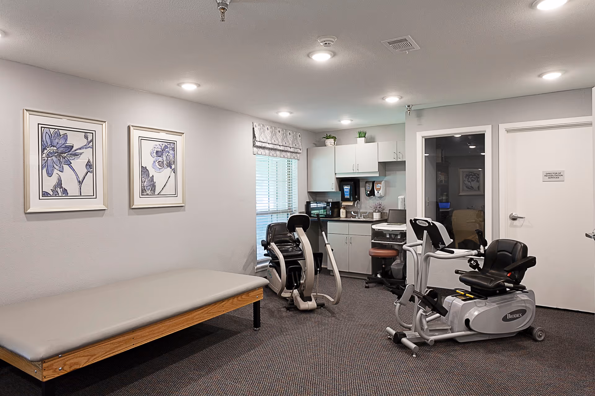 Small interior rehab room with exercise bikes, a therapy table, and cabinets.
