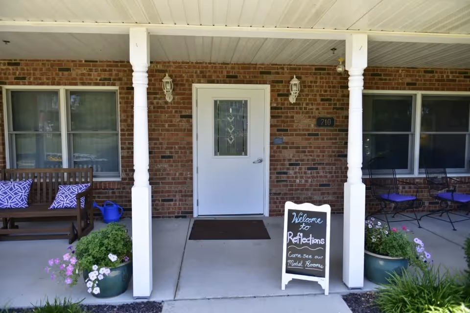 Front porch of a brick building with a white door in the center, flanked by two white columns. There are two windows on each side of the door. On the left side, there is a wooden bench with purple patterned cushions and a blue watering can. On the right side, there are two black metal chairs with purple cushions. Two large flower pots with green plants and flowers are placed near the columns. A small chalkboard sign near the door reads 'Welcome to Reflections Come see our Model Rooms.'