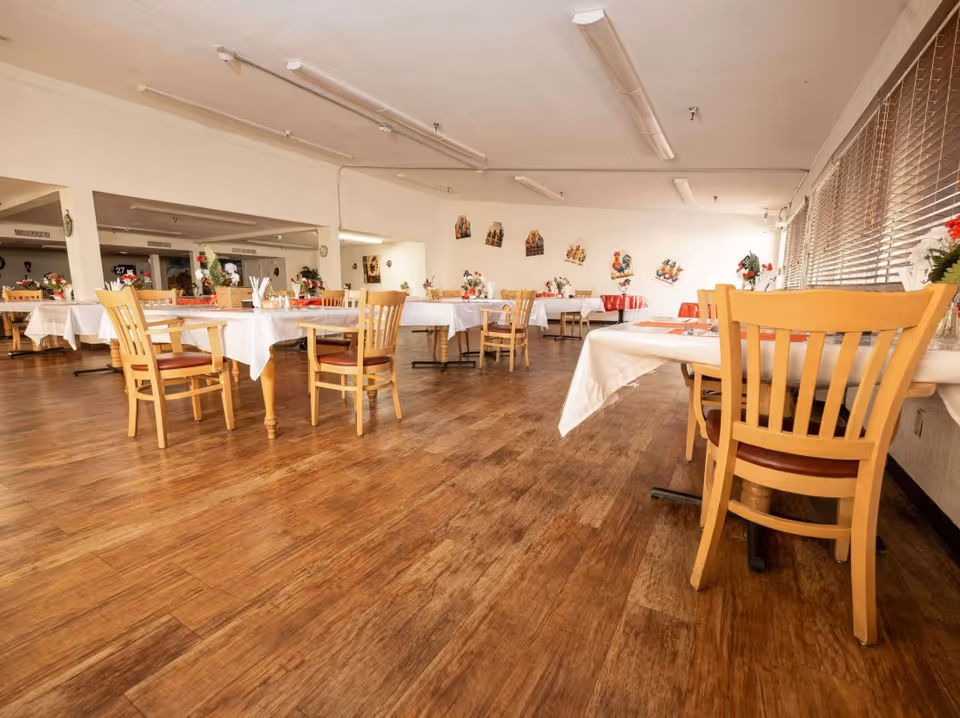 A spacious dining room with wooden floors and multiple tables covered with white tablecloths. Each table is surrounded by wooden chairs with red cushions. The room is decorated with small flower arrangements and wall art, and large windows with blinds allow natural light to enter.